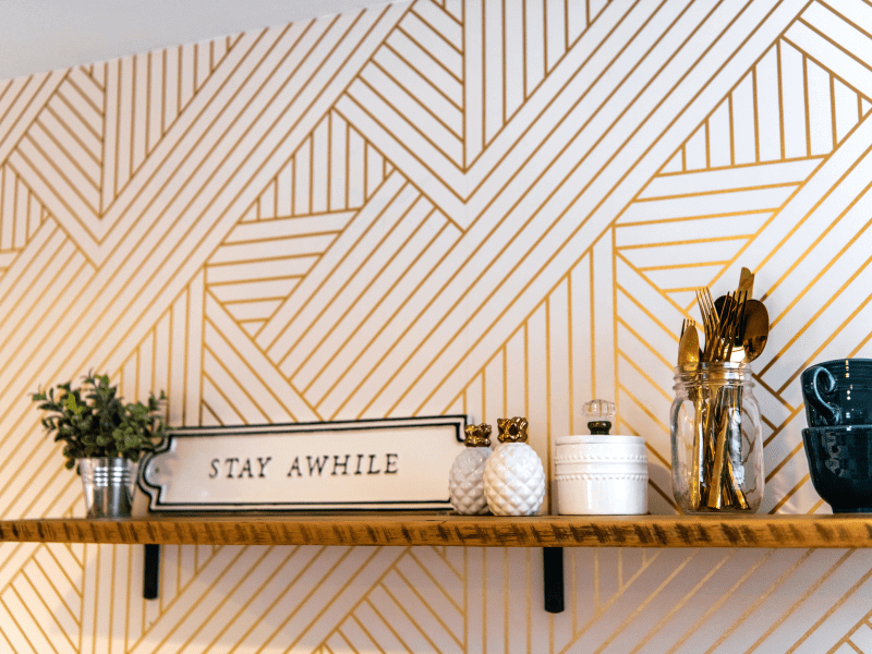 A geometrically designed wall with gold stripes, a shelf with a sign saying Stay awhile