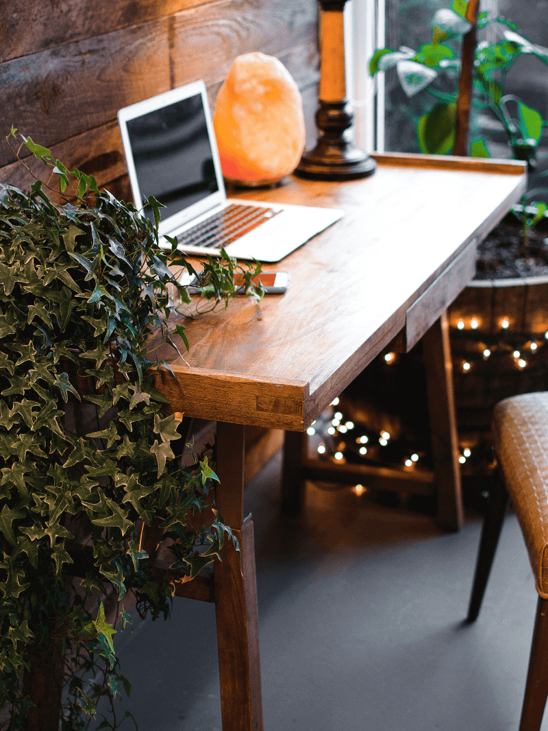 A desk with a laptop and salt lamp on it. Plants and fairy lights are around the desk